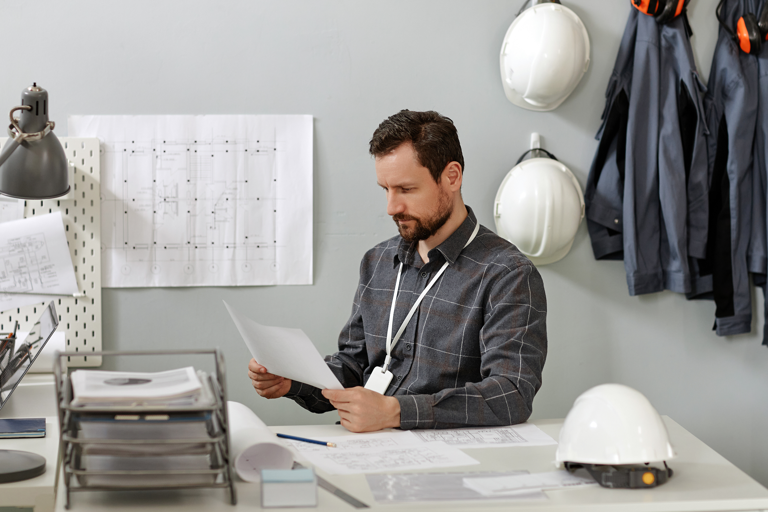 Man in an office reviewing HR paperwork at a desk covered with blueprints and documents, with white hard hats and work jackets hanging on the wall behind him.