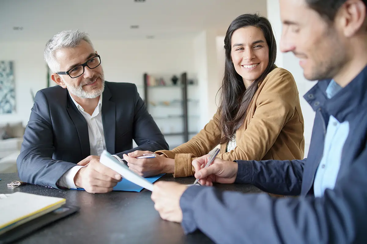 Three business professionals sit around a conference table reviewing documents during a meeting. A gray-haired man in a suit listens attentively while a woman smiles and another colleague reviews paperwork, creating a collaborative and professional workplace setting.