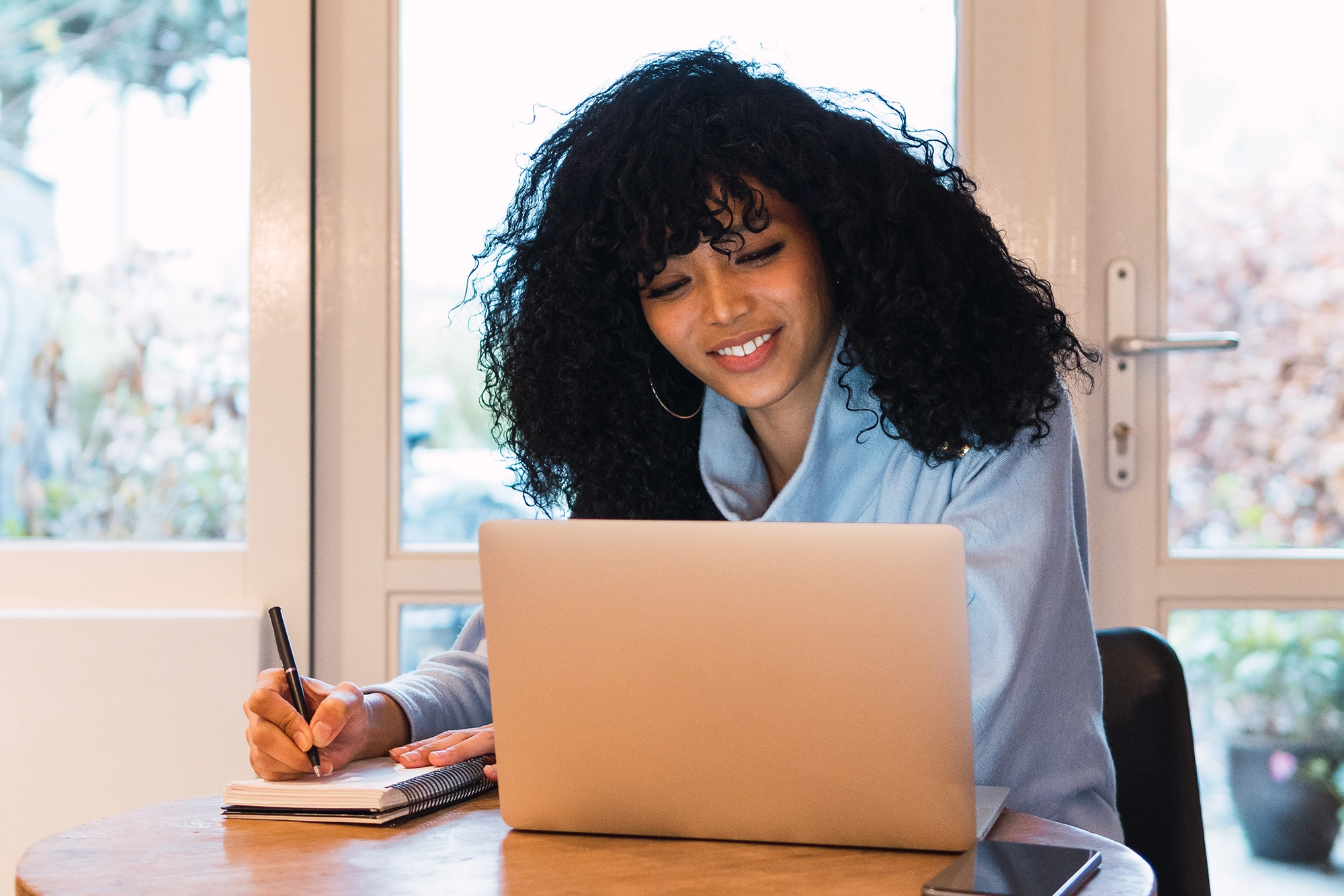 Young professional smiling while taking notes on a notebook beside a laptop, representing a positive employer brand and an engaging workplace that attracts job candidates.