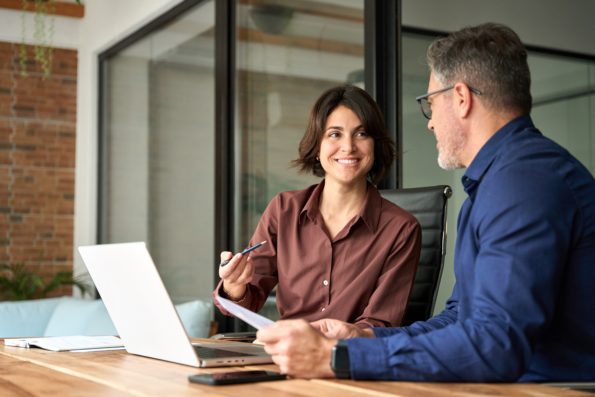 Two business professionals discussing work together at a desk, with a laptop and documents in a modern office.